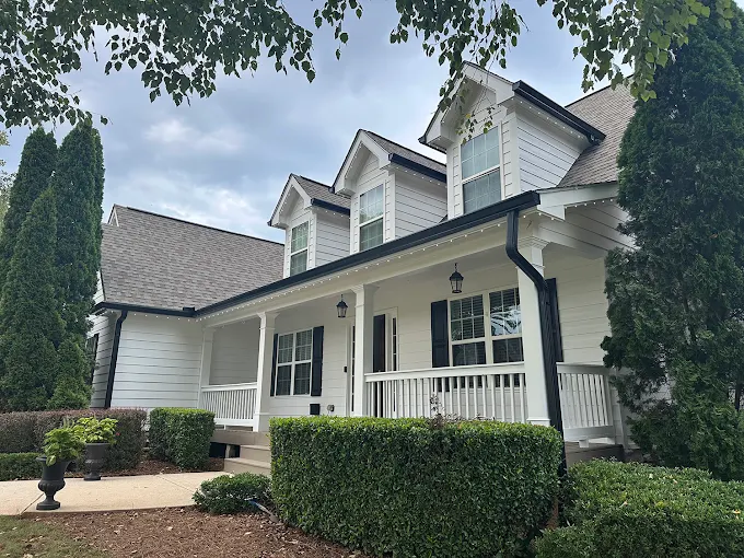 Two-story white house with black trim, front porch, dormer windows, and neatly trimmed bushes, set under a partly cloudy sky.