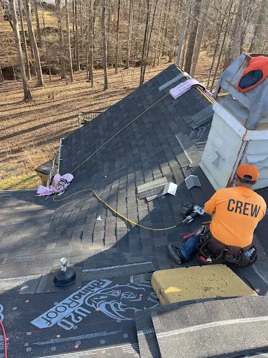 A roofer wearing an orange CREW shirt works on shingling a house roof surrounded by trees, with tools and roofing materials visible nearby.