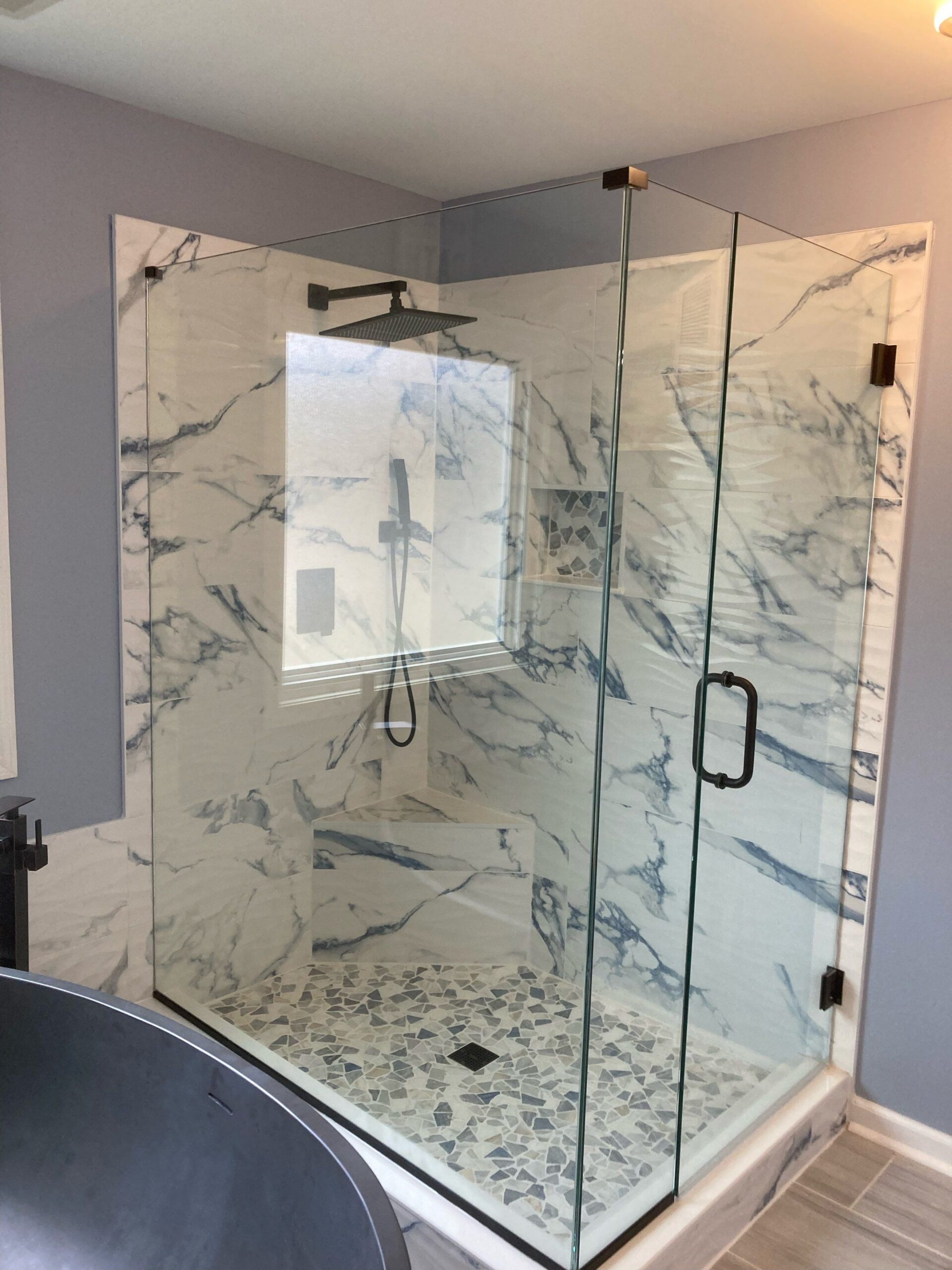 A modern walk-in shower with glass walls, marble-patterned tile, a built-in bench, pebble floor, and a wall-mounted showerhead.