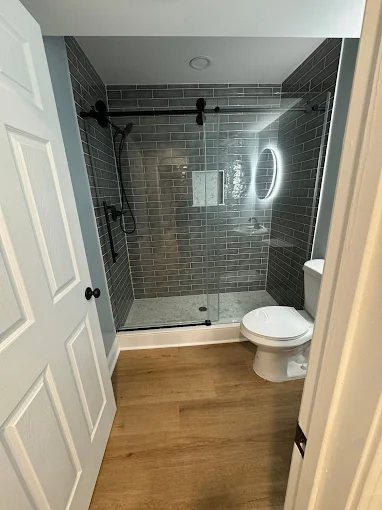 Modern bathroom with glass-enclosed shower, gray subway tile walls, circular LED mirror, white toilet, and light wood flooring.