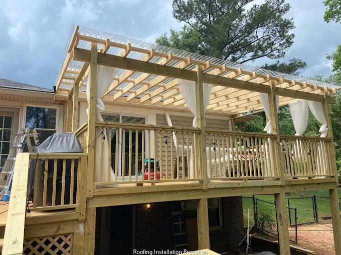 A wooden deck with a pergola roof, white curtains, and railings is attached to a house. There are outdoor furniture items and trees in the background.