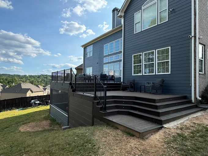 A modern house with blue siding features a raised wooden deck with black railings and outdoor seating, overlooking a grassy backyard and neighboring houses.