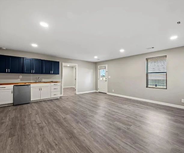 Unfurnished room with wood-style flooring, a kitchenette with blue and white cabinets, stainless steel dishwasher, window, and door leading to another area.
