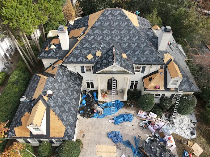 Aerial view of a large house undergoing roof repairs, with construction materials, tarps, and ladders positioned around the building.