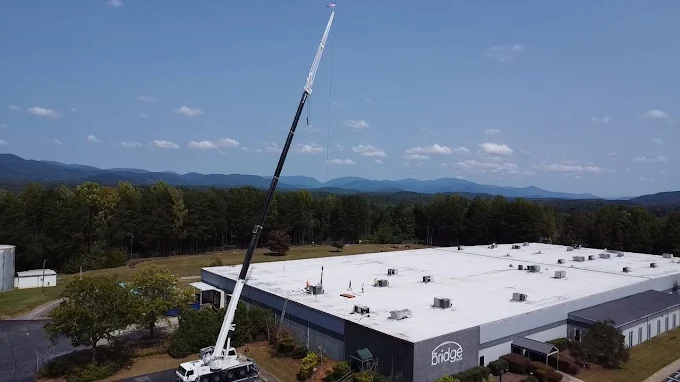 A large crane is parked next to a commercial building with a white roof, lifting or lowering materials; trees and mountains are visible in the background under a partly cloudy sky.